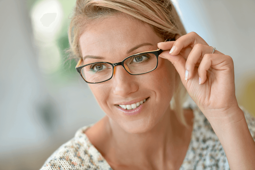 Attractive middle-aged woman with light hair and black-rimmed glasses, adjusting her eyewear with a gentle smile. A stylish choice in glasses for women over 50, highlighting both elegance and functionality.